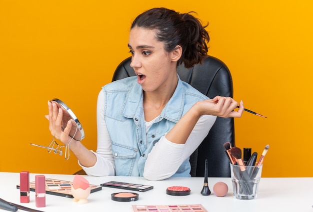Photo surprised pretty caucasian woman sitting at table with makeup tools holding makeup brush and looking at mirror