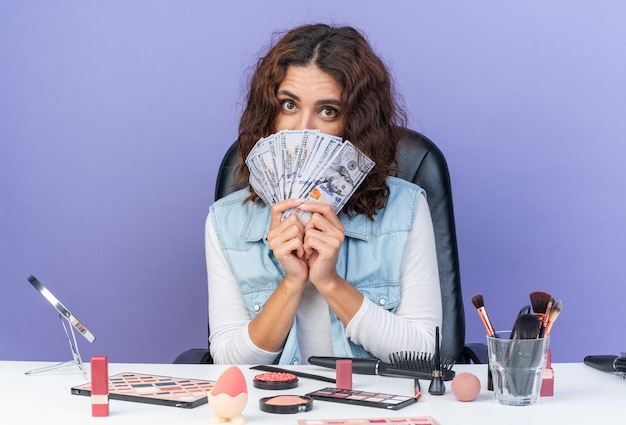 Surprised pretty caucasian woman sitting at table with makeup tools holding money isolated on purple wall with copy space