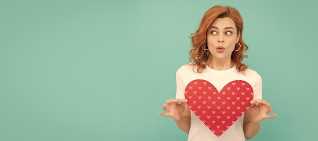 Photo surprised redhead girl hold red heart on blue background woman isolated face portrait banner with copy space