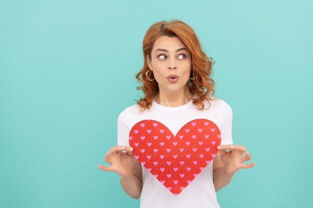 Surprised redhead girl hold red heart on blue background