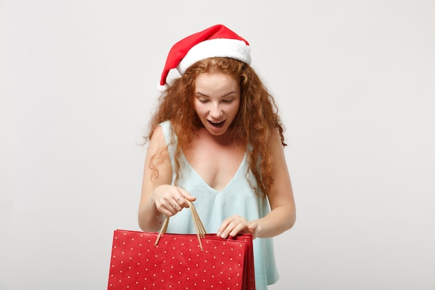 白い背景で隔離のクリスマス帽子で驚いた赤毛のサンタの女の子。明けましておめでとうございます2020年のお祝いの休日のコンセプト。コピースペースをモックアップします。買い物の後にギフトや購入品が入ったパッケージバッグを持ってください。