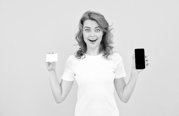 Удивленная рыжая женщина показывает кредитную или дебетовую карту и смартфон, чтобы совершать покупки в Интернете