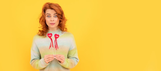 Surprised redhead woman with red heart sticks on yellow\
background woman isolated face portrait banner with copy space
