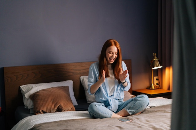 Surprised redhead young woman getting good online news on mobile phone at bedroom. Happy red-haired female enjoy success on cellphone at home sitting on bed with crossed legs at light living room.