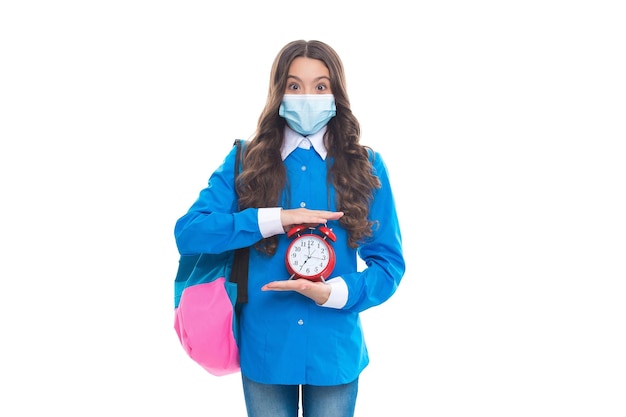 Surprised school child wear protective medical mask to prevent COVID19 disease spread holding alarm clock deadline