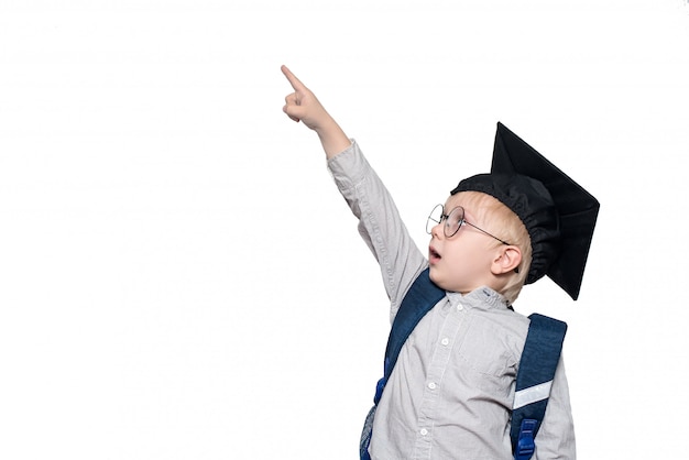 Surprised schoolboy in a suit, glasses and an academic hat points his finger up. School concept. Isolate