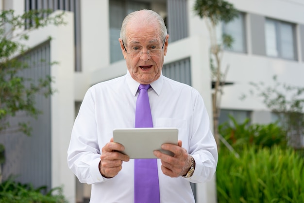 Surprised senior man in white shirt, tie and glasses looking at tablet screen outdoors.