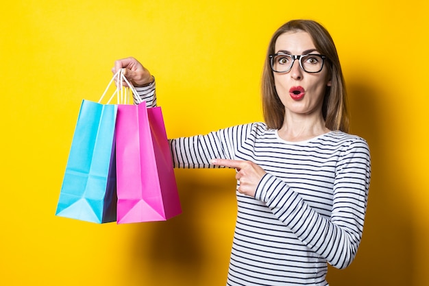 Surprised in shock, a young woman points a finger at shopping bags on a yellow background