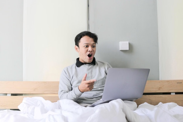 Surprised and shocked Asian man is working with his laptop on his cozy bed Concept of successful freelancer lifestyle