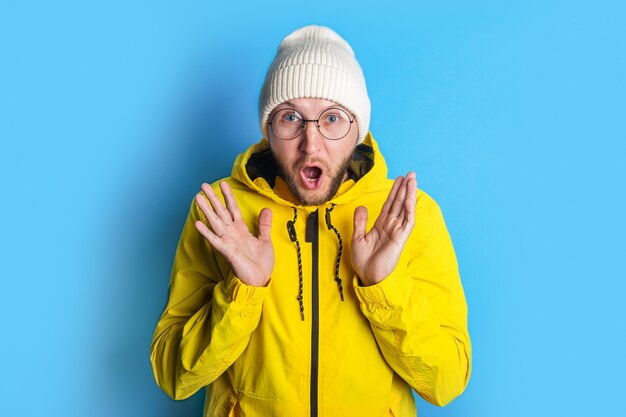 Surprised shocked bearded young man with glasses in a yellow jacket on a blue background.