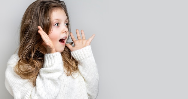 Surprised shocked little girl happily looks away and holds her hands to her face on a light background. Copy space, mock up