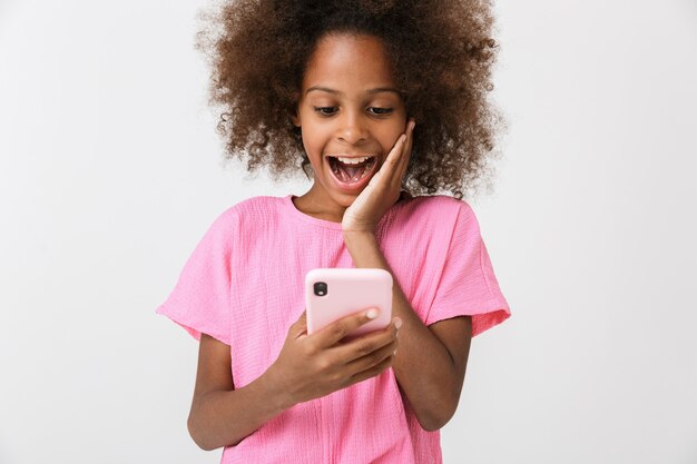 surprised shocked young african girl kid posing isolated over white wall, using mobile phone.