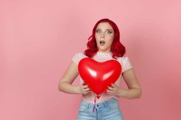 a surprised, shocked young woman with red hair holds a large flying red heart-shaped balloon