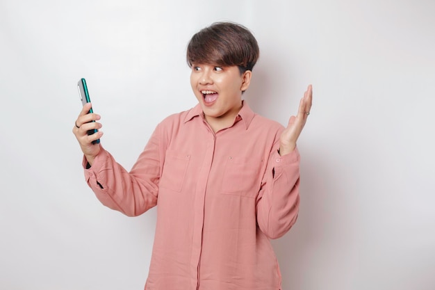 Surprised shorthaired Asian woman wearing a pink shirt pointing at her smartphone isolated by a white background