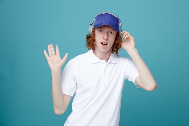 Surprised showing stop gesture young handsome guy in cap wearing headphones isolated on blue background
