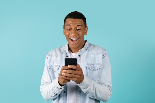 Surprised smiling young african american guy in casual typing on smartphone isolated on blue background