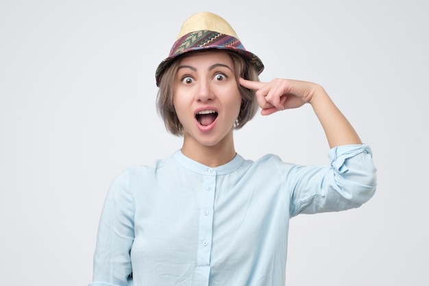 Surprised smiling young woman in summer hat twisting her index finger at temple