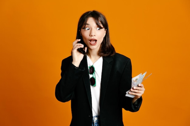 Surprised speaks on the phone holding money young beautiful female wearing black jacket isolated on orange background