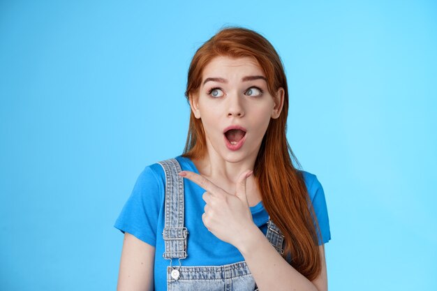 Surprised speechless ginger girl react shocked interested awesome copy space price fall, drop jaw pointing looking stunned left, open mouth fasinated amazed, stand blue background astonished