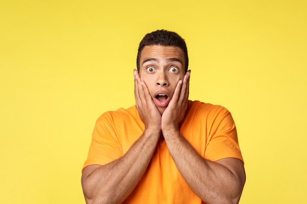 Surprised and speechless young shocked handsome man in t-shirt, gasping amazed, drop jaw and stare camera popped eyes, grab face from amazement, standing stupor from realisation, yellow background