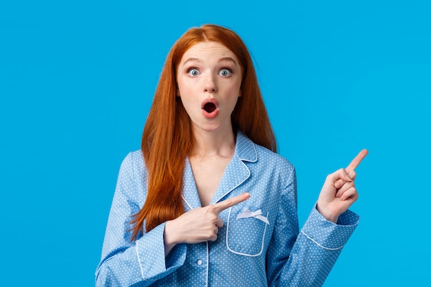 Surprised startled redhead girl found out special offer, discount promo, pointing upper right corner, wearing nightwear, gossiping about sleepover host, standing blue wall