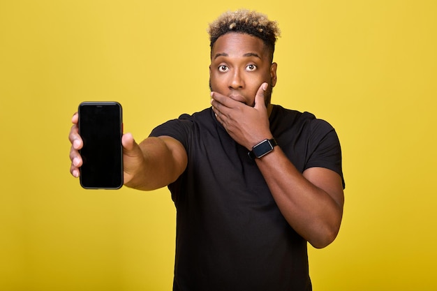 Surprised tall black man holds a mobile phone in his hand on yellow background