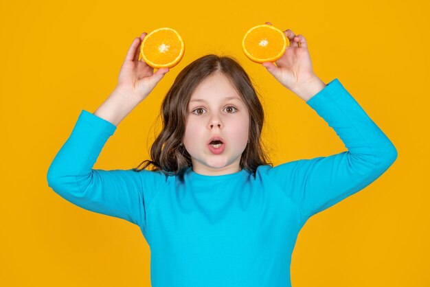 Surprised teen girl hold orange fruit on yellow background