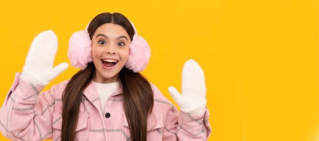 Surprised teen girl in winter mittens and earmuffs on yellow background winter fashion Banner of christmas child girl studio kid winter portrait with copy space