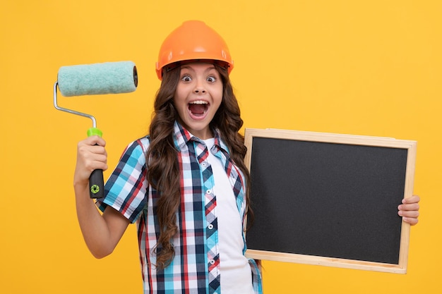 Surprised teen girl with curly hair in helmet hold paint roller and school blackboard copy space, education.