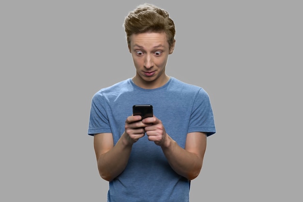 Surprised teenage boy using mobile phone. Trendy teen guy using app on his smartphone against gray background.