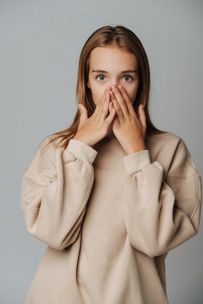 Surprised teenage girl covering her face with hands, looking at the camera. Against grey background. In a warm beige longsleeve.