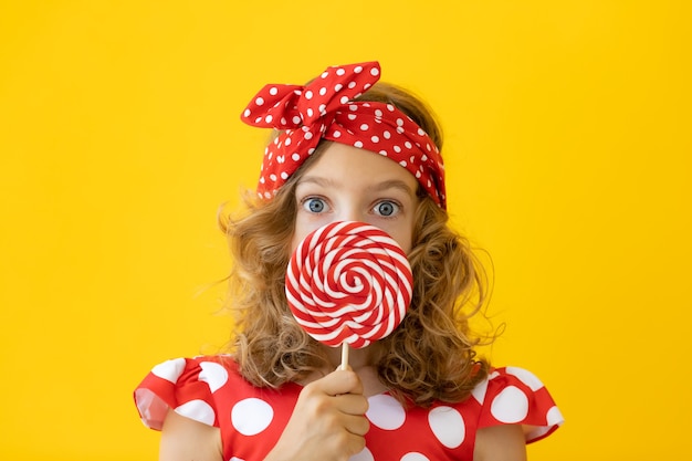 Surprised teenager girl holding red lollipop against yellow wall