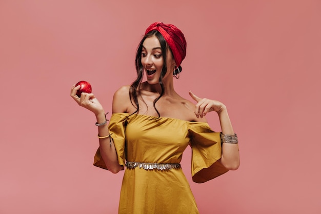 Surprised wavy haired trendy lady with cool hairstyle in red bandana and bright sundress looking at apple on isolated pink backdrop