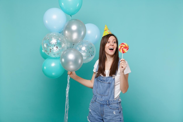 Surprised woman in birthday hat keeping mouth open, hold round lollipop, celebrating with colorful air balloons isolated on blue turquoise background. Birthday holiday party, people emotions concept.