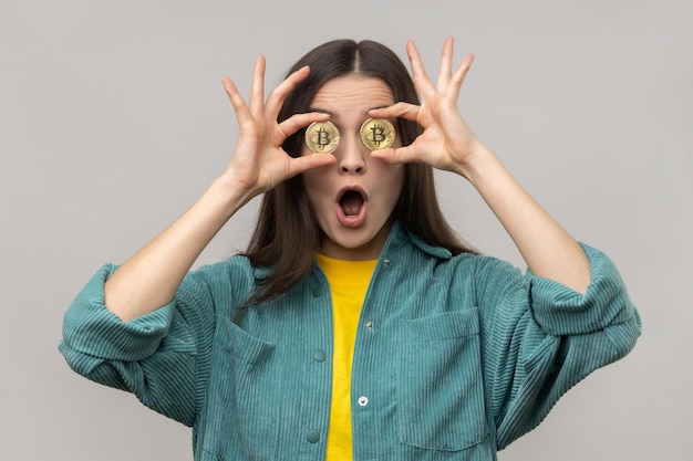 Surprised woman covering eyes with golden bitcoin looking camera through btc coin keeps mouth open