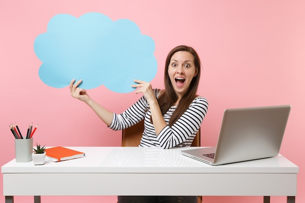 Surprised woman hold blue empty blank Say cloud speech bubble work at white desk with pc laptop isolated on pastel pink background. Achievement business career concept. Copy space for advertisement.