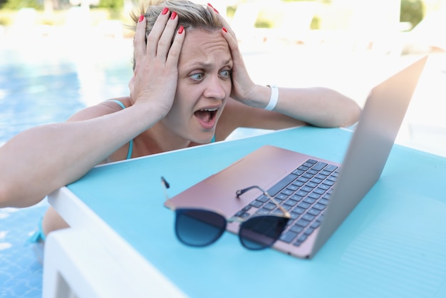 Surprised woman holding her head and looking at laptop screen on side of pool