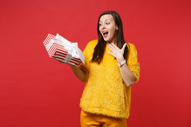 Surprised woman keeping mouth open, hold red striped present box with gift ribbon isolated on red background. Valentine's Day, International Women's Day, birthday, holiday concept. Mock up copy space.