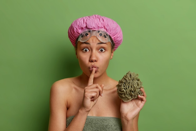 Surprised woman makes shush gesture tells secret holds bath sponge waterproof hat and goggles going to take shower wrapped in towel isolated over green studio wall