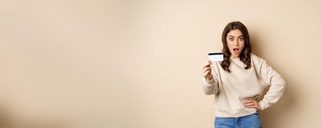 Surprised woman showing credit card gasping impressed impression of discounts or shopping standing over beige background