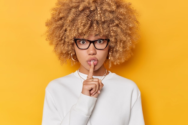 Surprised woman with curly bushy hair makes silence gesture keeps index finger over lips looks mysteriously wears transparent glasses and casual white jumper isolated over yellow background