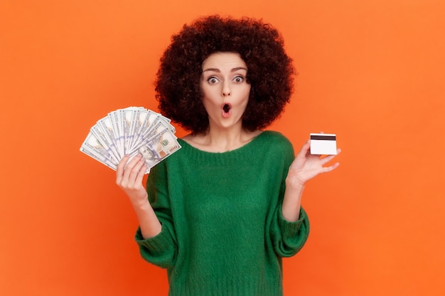 Surprised woman with curly hair in green sweater holding fan of dollars banknotes and credit card, benefits of bank loan, pondering deposit percentage. Indoor studio shot isolated on orange background