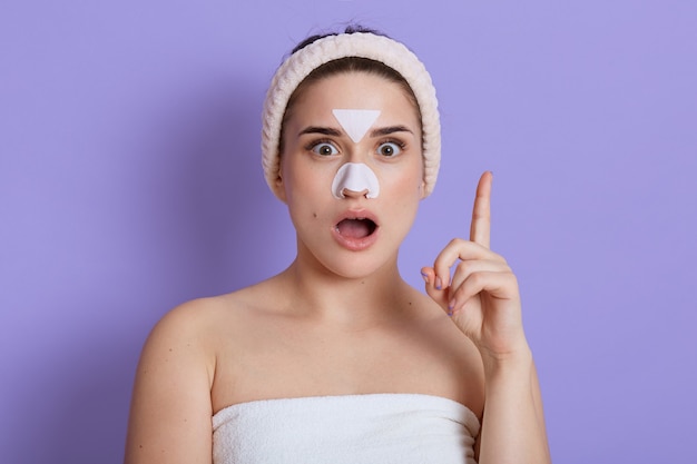 surprised woman with facial mask on nose and forehead, has beauty procedures, shocked expression, points up with index finger, wrapped towel and wears hair band, isolated over lilac wall.