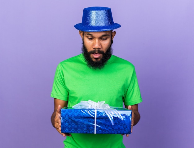 Удивленный молодой афро-американский парень в партийной шляпе, держащий и смотрящий на подарочную коробку, изолированную на синей стене