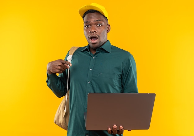 Surprised young afro-american student with cap and backpack holding and pointing at laptop isolated on orange wall with copy space