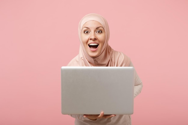 Surprised young arabian muslim woman in hijab light clothes posing isolated on pink background. People religious Islam lifestyle concept. Mock up copy space. Holding and working on laptop pc computer.