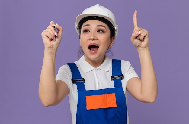 Giovane ragazza asiatica sorpresa del costruttore con il casco di sicurezza bianco che esamina l'indicatore e che indica su