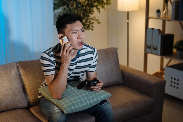 Surprised young asian chinese man holding joystick and looking\
desperate while sitting on couch at home in night. guy calling\
friend on cellphone asking for help to win video game in\
evening.