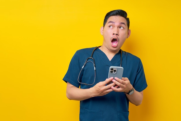 Surprised young asian male doctor or nurse wearing blue uniform with open mouth and holding mobile phone while looking aside isolated on yellow background healthcare medicine concept