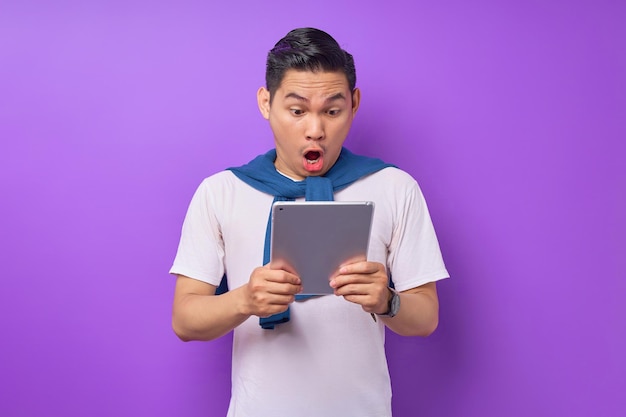 Surprised young Asian man in casual clothes holding a digital tablet and open mouth isolated over purple background